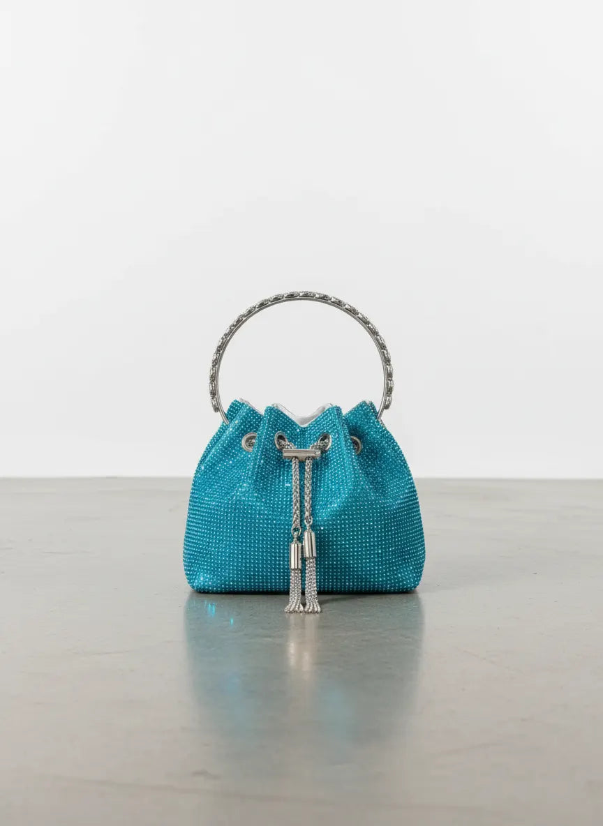 Turquoise woven handbag with silver handle and tassel on a reflective surface.